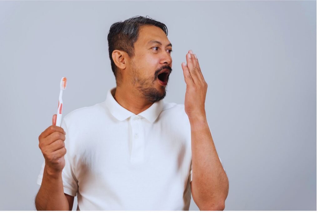 Photo of a man holding a toothbrush with one hand and covering his mouth because of bad breath with his other hand.