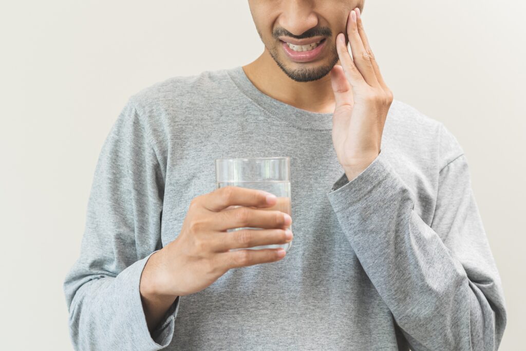 Photo of a main drinking a cold glass of water and grimacing in pain from sensitive teeth.